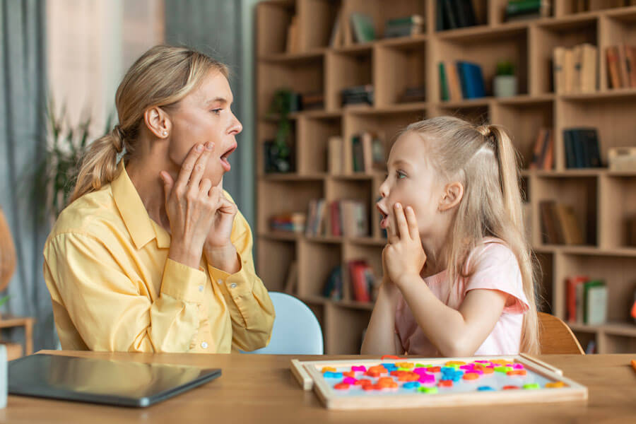 A caucasian woman sits at a table and faces her left with her mouth open and her fingers positioned on her cheeks. A young caucasian girl sits opposite and faces her, mirroring the woman's actions.