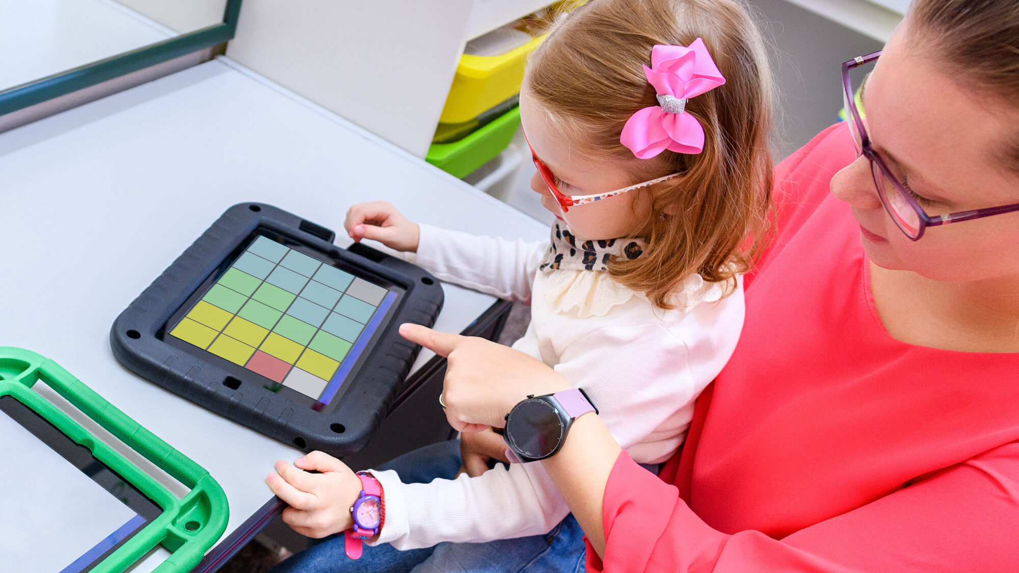 A young girl sits on a woman's lap as at a desk and they look at an Augmentative and Alternative Communication device.