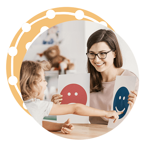 A young caucasian woman with long, straight brown hair and wearing glasses sits at a table opposite a a very young girl. The woman smiles and holds up a picture of a frowning face in her right hand and a smiling face in her left hand as the young girl points to the smiling face.